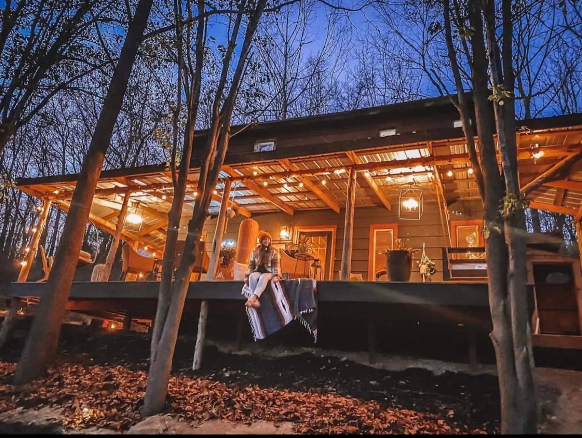 The cabin glowing at dusk, its wraparound porch strung with lights and framed by bare Ozark trees